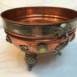 May include: A copper bowl with a silver rim and decorative accents. The bowl has three ornate feet and a handle with a decorative face. The bowl is decorated with small, colourful stones.