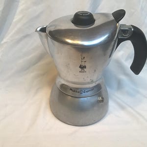May include: A stainless steel Moka Express coffee maker with a black handle and lid knob. The coffee maker has the Bialetti logo and the words "Mukka Express" printed on it. The coffee maker is designed to brew espresso.