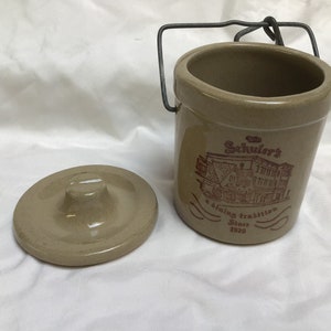 May include: A beige ceramic crock with a metal handle and lid. The crock has a brown and white image of a building with the text "The Schuler's A dining tradition Since 1928".