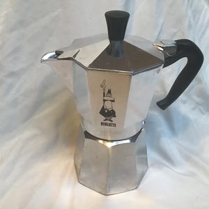 May include: A silver stovetop espresso maker with a black handle and top. The coffee pot has an octagonal base and a black and white cartoon figure on the front, with the word "BIALETTI" below.