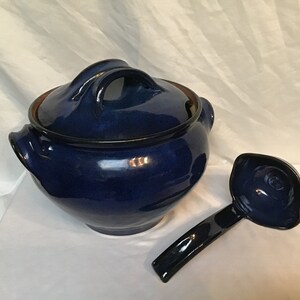 Op de afbeelding: Een donkerblauwe keramische soepterrine met bijpassend deksel en pollepel. De terrine heeft een afgeronde vorm met twee handvatten en een donkere rand. De pollepel heeft een lange steel en een afgeronde kom. De set staat op een wit oppervlak.