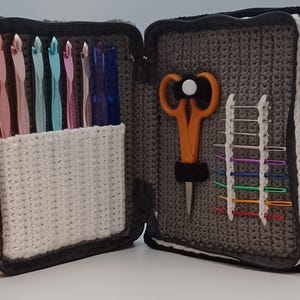 Puede incluir: Estuche abierto para ganchillos con varios ganchillos de colores, tijeras naranjas y marcadores de punto coloridos. El estuche es gris con un bolsillo de ganchillo blanco y ribete negro. El estuche está abierto, mostrando el contenido.