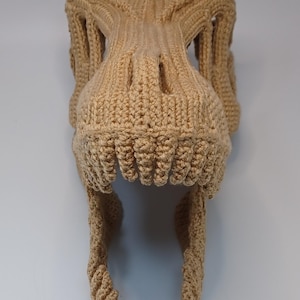 May include: A tan-colored, crocheted dinosaur skull, featuring detailed teeth and eye sockets. The handmade piece is likely a costume or decorative item, showcasing a textured, three-dimensional design.