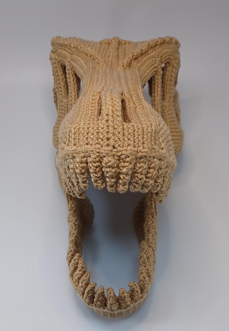 May include: A tan-colored, crocheted dinosaur skull, featuring detailed teeth and eye sockets. The handmade piece is likely a costume or decorative item, showcasing a textured, three-dimensional design.