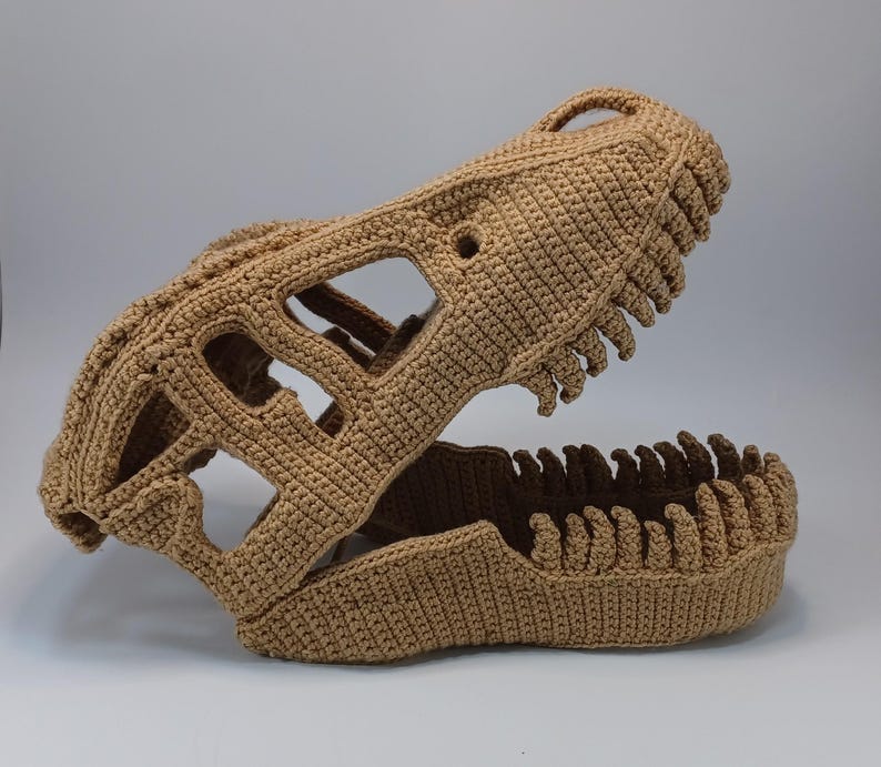 May include: A crocheted dinosaur skull in a tan color. The skull features open eye sockets and a detailed jawline with teeth. The handmade piece is displayed against a plain background, showcasing the intricate crochet work.