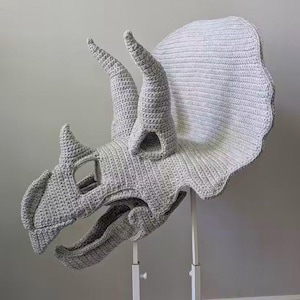 May include: A light grey crochet triceratops dinosaur head mount featuring a large frill, two prominent brow horns, and a smaller nose horn. The textured yarn construction displays visible stitches throughout the entire dinosaur skull form.