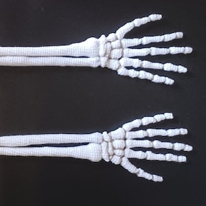 Könnte beinhalten: Zwei weiße gehäkelte Arme mit Händen, die einem Skelett ähneln. Die Arme befinden sich auf einem schwarzen Hintergrund.