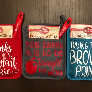 Pode incluir: Três luvas de forno vermelhas, cinzas e azuis com bolsos. Cada luva tem um pacote de mistura de chocolate ou brownie Betty Crocker e uma espátula vermelha dentro do bolso. As luvas têm diferentes dizeres: "obrigado por me fazer um biscoito inteligente", "como é doce ser ensinado por você", e "tentando ganhar pontos brownie".