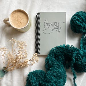 Peut inclure: Un journal de projet à spirales gris clair avec du texte noir, une tasse de café, de la laine vert d'eau et des fleurs séchées sur une surface blanche. Le journal est ouvert, et le texte est manuscrit.