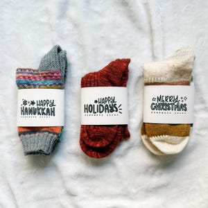 May include: Three pairs of handmade socks, each wrapped with a white band featuring holiday greetings. The socks are in shades of gray, red, and cream, with colorful stripes and patterns. The text on the bands reads "Happy Hanukkah", "Happy Holidays", and "Merry Christmas".