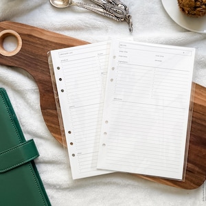 May include: A wooden chopping board with a handle holds two white paper inserts with lined pages and the words "Grocery List" and "Recipe". A green binder is in the lower left corner. A spoon and a muffin are in the background.