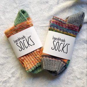 Könnte beinhalten: Zwei Paar handgemachte Socken mit weißen Etiketten. Eine Socke ist orange, grün und gelb. Die andere ist grau, blau und rot. Auf den Etiketten steht "handmade SOCKS" in schwarzer Schrift.