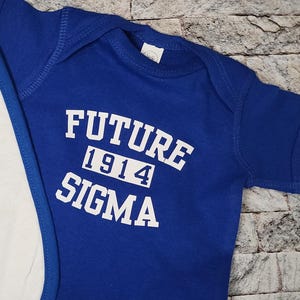 May include: A royal blue baby onesie with the words "FUTURE 1914 SIGMA" printed in white. The onesie is made of soft material and has short sleeves. A white bib with a blue trim is partially visible.