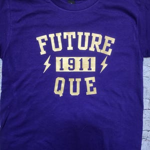 May include: Purple t-shirt with gold lettering that reads "FUTURE 1911 QUE".