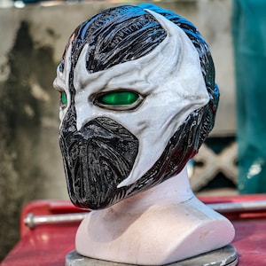 May include: A full-head mask featuring a high-contrast design with glossy black textured sections and smooth white accents. The mask includes striking, vibrant green eye lenses and a detailed, moulded facial structure covering the entire head.