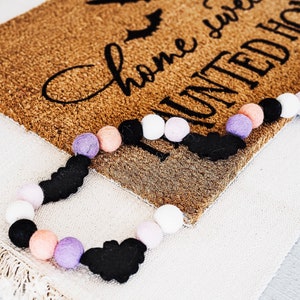 Puede incluir: Una alfombra de bienvenida marr&oacute;n con el texto "Home sweet haunted home" en letras negras. Una guirnalda de murci&eacute;lagos de fieltro negro y pompones de color rosa pastel, morado y blanco est&aacute; drapeada sobre la alfombra.
