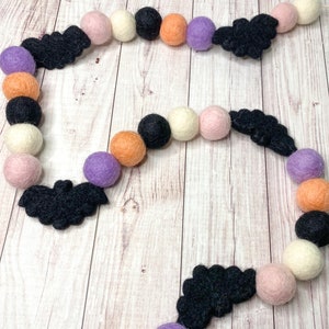 Puede incluir: Una guirnalda de Halloween con murci&eacute;lagos de fieltro negro, bolas de fieltro naranja, blanco, rosa y morado.