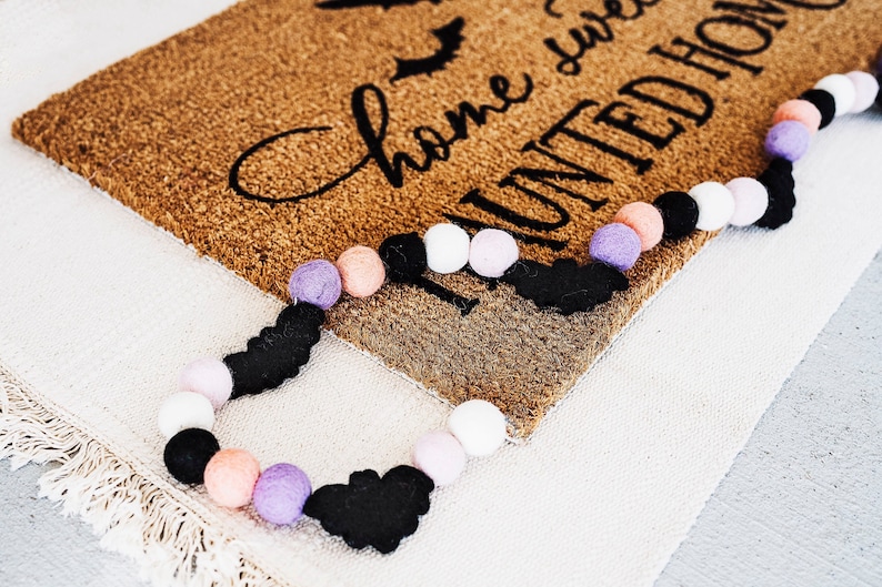 Puede incluir: Una alfombra de bienvenida marr&oacute;n con el texto "Home sweet haunted home" en letras negras. Una guirnalda de murci&eacute;lagos de fieltro negro y pompones de color rosa pastel, morado y blanco est&aacute; drapeada sobre la alfombra.