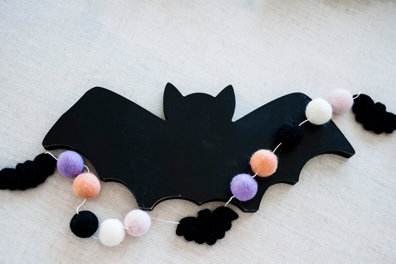 Puede incluir: Un murci&eacute;lago de fieltro negro con una cadena de bolas de fieltro en morado, rosa, blanco y naranja. Las bolas de fieltro est&aacute;n enhebradas con un hilo blanco y cuelgan de las alas del murci&eacute;lago.