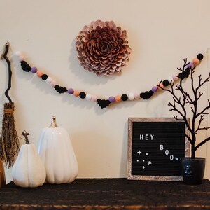 Puede incluir: Una guirnalda de Halloween con murci&eacute;lagos negros y pompones rosas, morados y blancos cuelga de una pared. Una calabaza de cer&aacute;mica blanca se encuentra sobre una mesa con un tablero de letras negro que dice "HEY BOO".