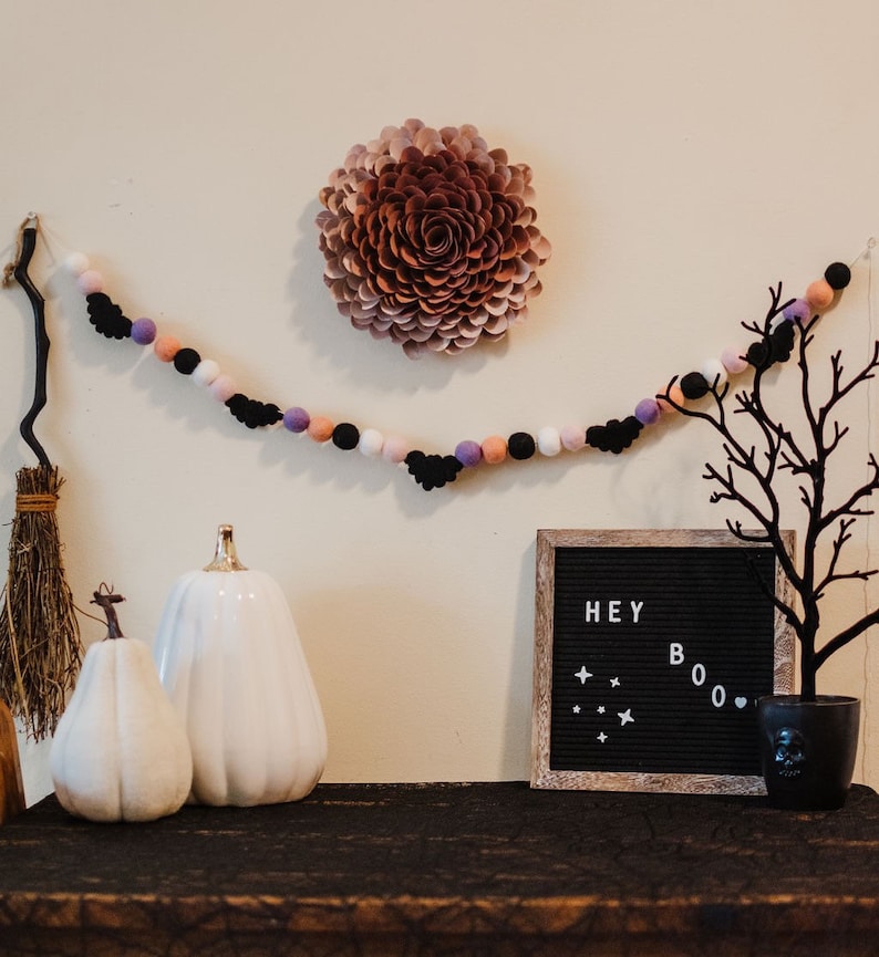 Puede incluir: Una guirnalda de Halloween con murci&eacute;lagos negros y pompones rosas, morados y blancos cuelga de una pared. Una calabaza de cer&aacute;mica blanca se encuentra sobre una mesa con un tablero de letras negro que dice "HEY BOO".