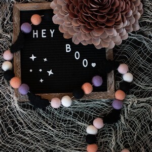 Puede incluir: Un tablero de letras negro con las palabras "HEY BOO" deletreadas en letras blancas. Una guirlande de bolas de fieltro negras, blancas, rosas y moradas est&aacute; drapeada sobre el tablero de letras y una gran flor de papel marr&oacute;n est&aacute; en el fondo.