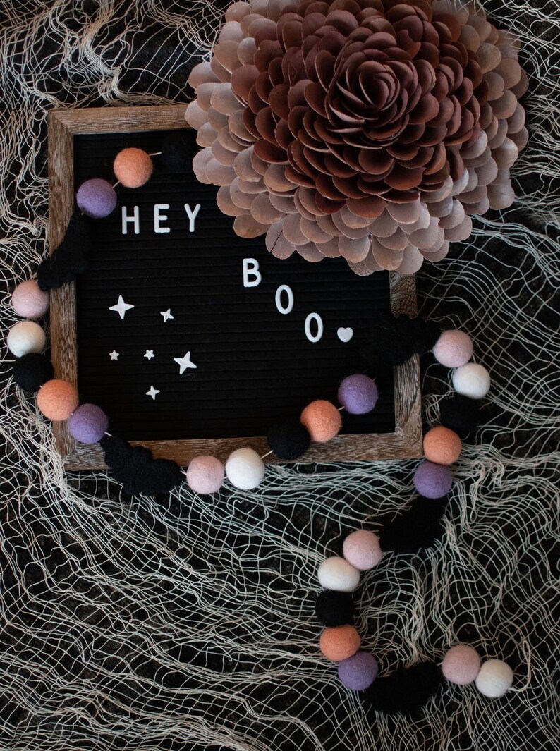 Puede incluir: Un tablero de letras negro con las palabras "HEY BOO" deletreadas en letras blancas. Una guirlande de bolas de fieltro negras, blancas, rosas y moradas est&aacute; drapeada sobre el tablero de letras y una gran flor de papel marr&oacute;n est&aacute; en el fondo.
