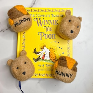 May include: Four felt toys shaped like Winnie the Pooh characters. The toys are brown and yellow and have embroidered details. The toys are arranged around a copy of "The Complete Tales of Winnie the Pooh" by A.A. Milne. The book has a yellow cover with black text.