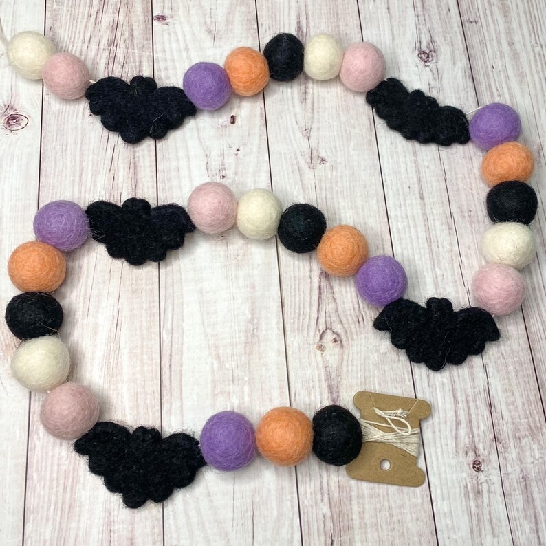 Puede incluir: Una guirnalda de Halloween con murci&eacute;lagos de fieltro negro y bolas de fieltro naranja, rosa, blanco y morado. La guirnalda est&aacute; enhebrada en un cordel blanco.