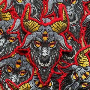 May include: A close-up of several embroidered patches featuring a three-eyed goat head with horns, red eyes, and a black beard. The patches are stitched with red, black, and gold thread on a grey fabric background.