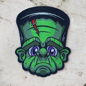 Può includere: Volto verde di Frankenstein con una cicatrice cucita sulla fronte, occhi grandi e un'espressione accigliata. Il mostro è in stile cartone animato e ha una linea di capelli nera.