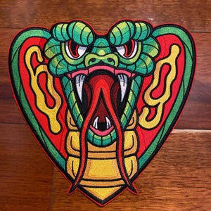 May include: A green, red, and yellow embroidered patch of a cobra snake with its mouth open and fangs showing. The snake is in a heart shape.