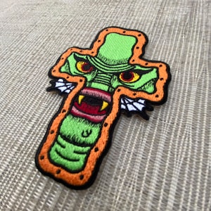 Könnte beinhalten: Grün-orangefarbener gestickter Aufnäher eines Monsters mit Zähnen und roten Augen auf einer Kreuzform.
