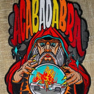 Puede incluir: Parche bordado con un mago con una túnica roja y la palabra "ACABADABRA" en letras coloridas. El mago sostiene una bola de cristal con un coche de policía en llamas. El parche tiene un borde negro.