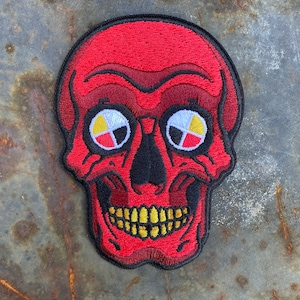 Può includere: Toppa di teschio ricamata rossa con occhi neri e bianchi che presentano un design di ruota di medicina nativo americano. Il teschio ha un ghigno dentato con denti gialli.