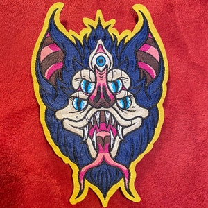 May include: A blue and pink embroidered patch of a bat with a third eye and multiple eyes on its face. The bat has a large mouth with sharp teeth and a long pink tongue.