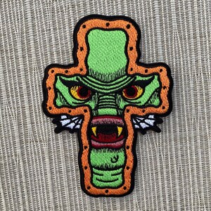 Peut inclure: Patch brod&eacute; vert repr&eacute;sentant un monstre aux yeux rouges et une grande bouche, en forme de croix avec un contour orange.