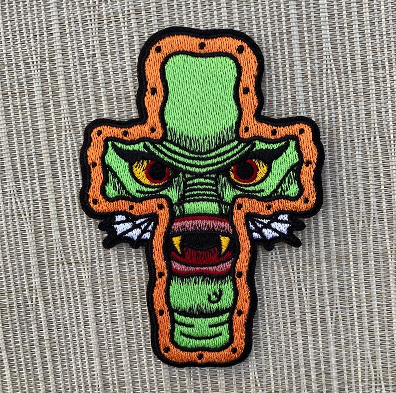 Peut inclure: Patch brod&eacute; vert repr&eacute;sentant un monstre aux yeux rouges et une grande bouche, en forme de croix avec un contour orange.