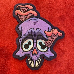 Könnte beinhalten: Lilafarbener Schädel mit einem rosa Pilz, der aus der Oberseite wächst. Der Schädel hat große, runde Augen mit blauen Pupillen. Der Pilz hat einen rosa Stiel und einen weißen Hut.