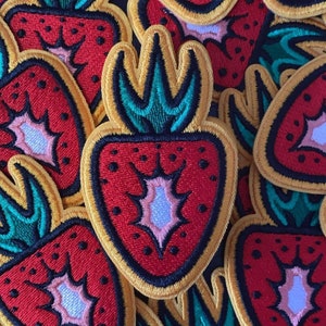 May include: A close-up of several embroidered iron-on patches featuring a red strawberry with a green stem and yellow outline. The strawberries have a white centre with a black starburst design.