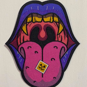 May include: A black and purple embroidered patch depicting a large mouth with sharp teeth and a pink tongue sticking out. The tongue has a small yellow square with a jack-o-lantern face on it.