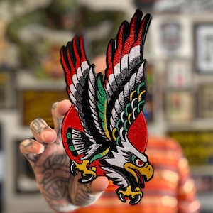 May include: An embroidered patch featuring a black and white eagle with green and yellow accents, flying against a red sun.