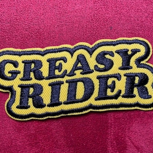 Peut inclure: Un patch brodé jaune et noir avec le texte "GREASY RIDER" dans une police rétro.