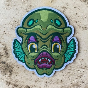 May include: Green and purple embroidered patch of a cartoon creature with large eyes and sharp teeth. The creature has a green and blue helmet and green fins.