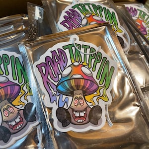 Könnte beinhalten: Ein Auto-Lufterfrischer mit einem psychedelischen Pilz-Design. Der Pilz ist bunt und hat ein lächelndes Gesicht. Der Text "Road Trippin'" ist in einer bunten, stilisierten Schrift um den Pilz herum geschrieben.