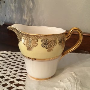 May include: A yellow and white creamer with a gold trim and a gold floral design. The creamer has a handle and a spout.