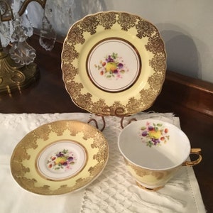 May include: A yellow teacup and saucer set with a gold trim and a floral design featuring a cluster of fruit. The set is on a white lace tablecloth.