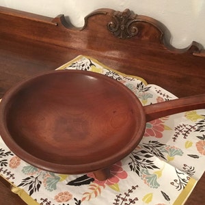 May include: A wooden bowl with a handle and three legs. The bowl is brown and has a smooth finish. It is sitting on a white and floral patterned napkin.