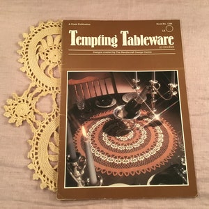 May include: A crochet pattern book titled "Tempting Tableware in crochet" with a photo of a crocheted table runner on a table setting with candles, a bottle of champagne, and a glass.