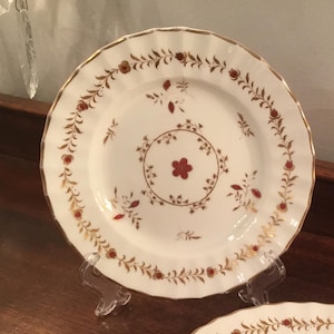 Puede incluir: Un plato de porcelana blanca con un diseño floral dorado y rojo. El plato tiene un borde festoneado y está sobre un soporte transparente.